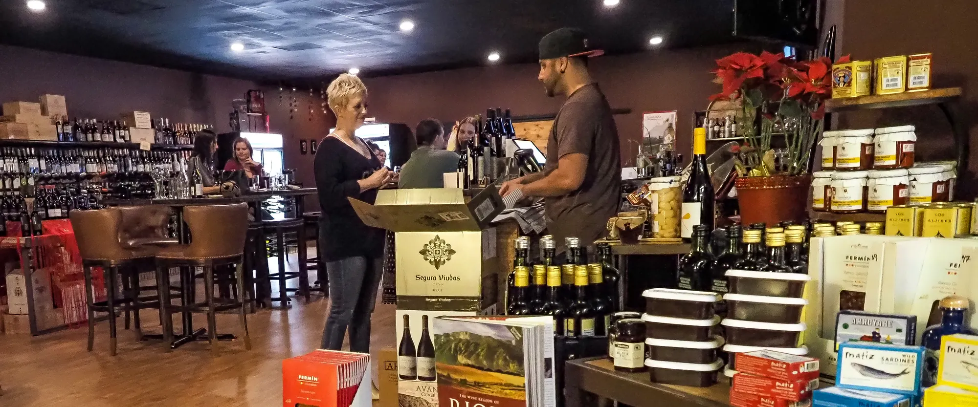 Inside La Vid: guests at the register, leather bar stools, wine walls along both sides, and crates, jamón, and Rioja books stacked on the floor