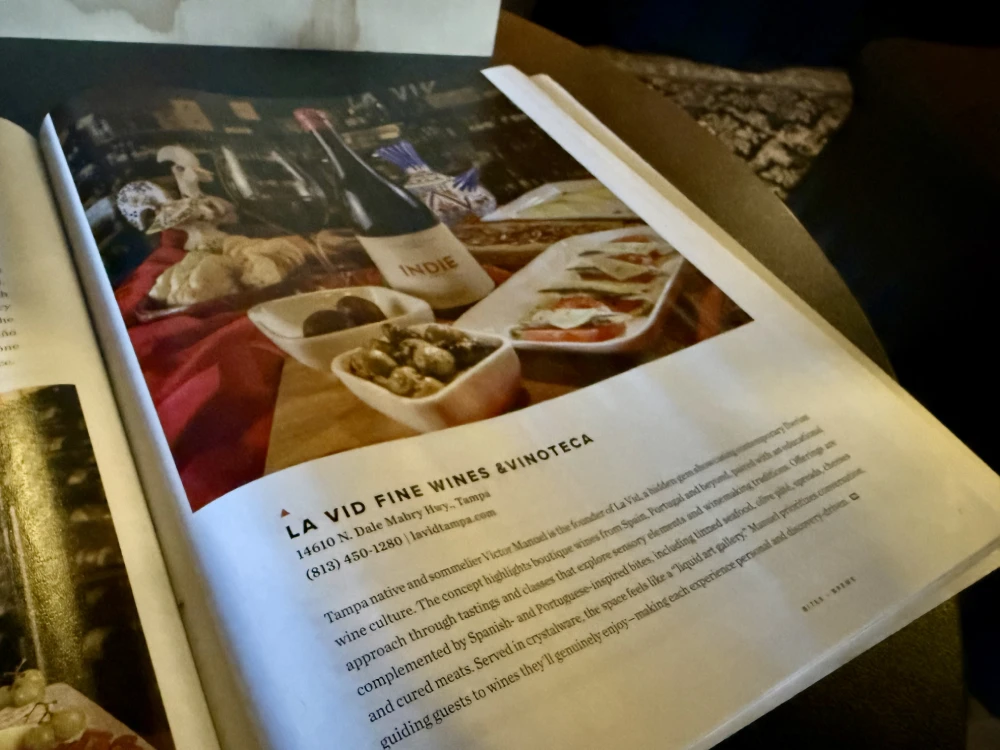 A printed guide open to a full-page feature on La Vid Fine Wines & Vinoteca, showing the shop's address and phone number beside a photograph of wine and small plates