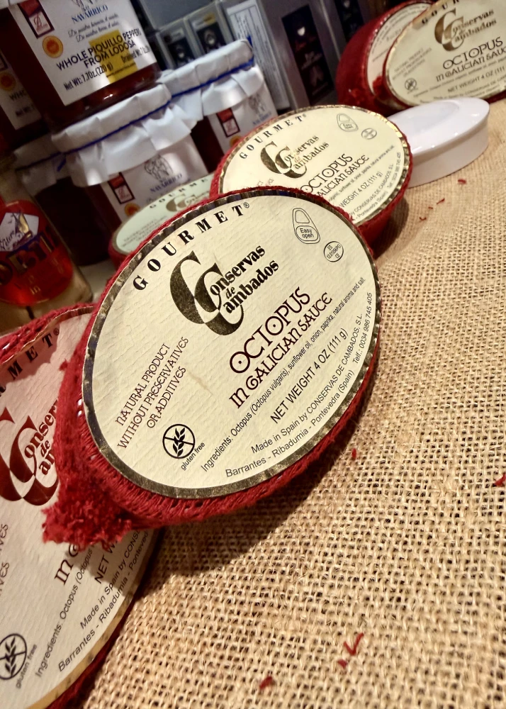 Tins of Conservas de Cambados octopus in Galician sauce, resting on burlap beside jars of piquillo peppers
