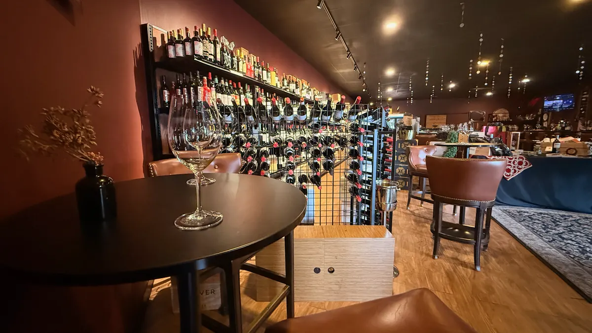 A small round table set with a wine glass beside a rack of bottles, the room’s tables and wine walls beyond