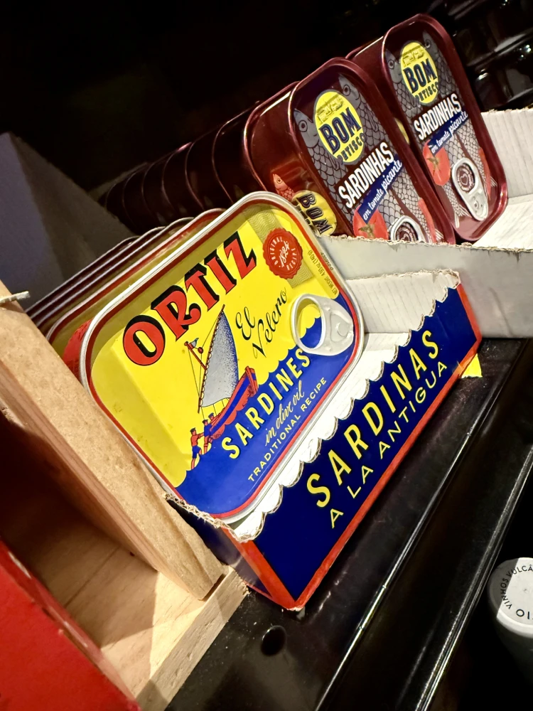 A tin of Ortiz El Velero sardines in olive oil in a wooden crate, with tins of Bom Petisco sardinhas behind it