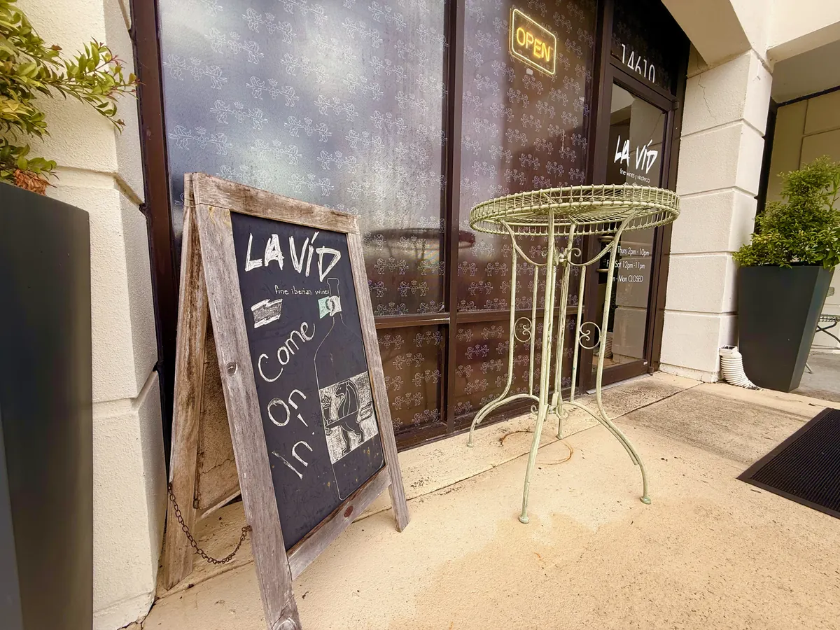 La Vid’s front door: an A-frame chalkboard reading “La Vid, come on in” and a wrought-iron bistro table beside the tinted windows