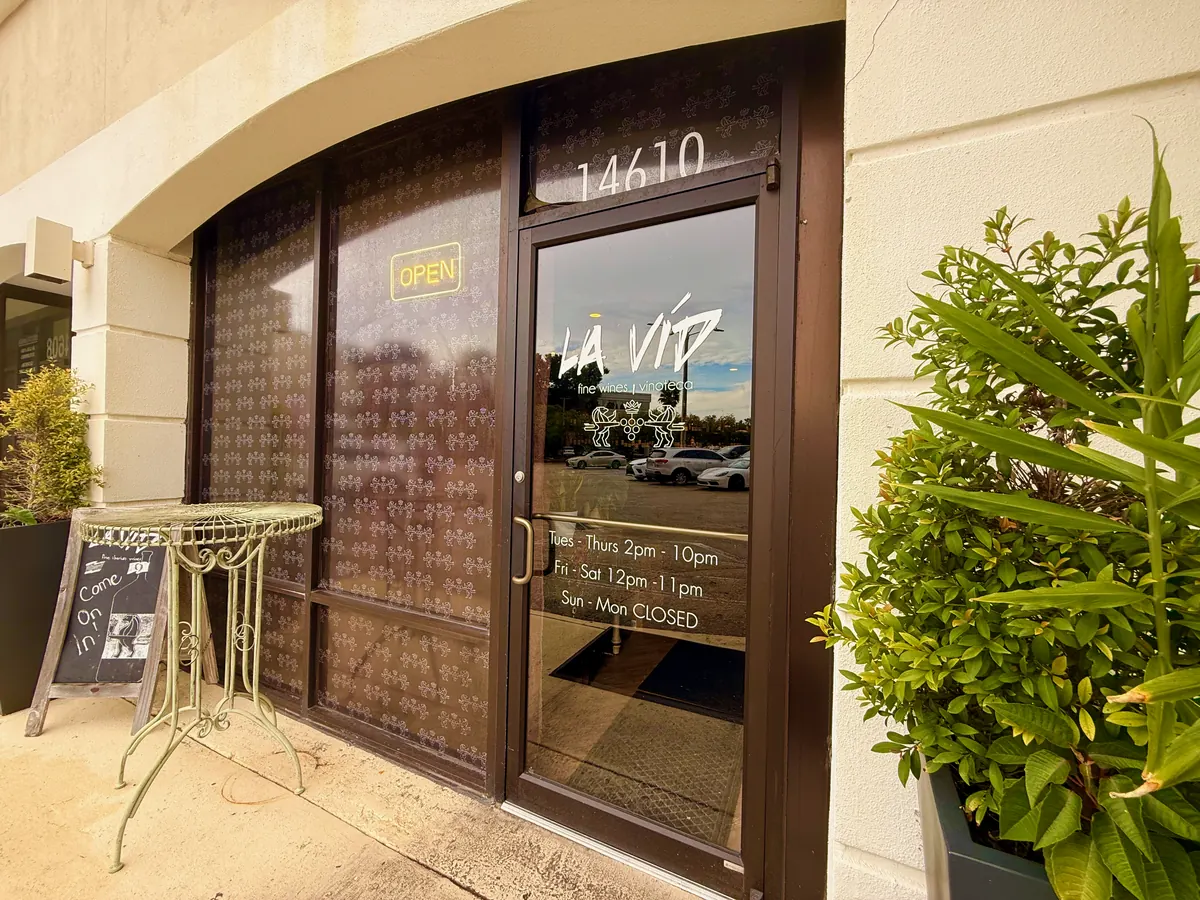 La Vid’s glass door under the 14610 address, with the La Vid logo and opening hours on the glass and potted plants either side